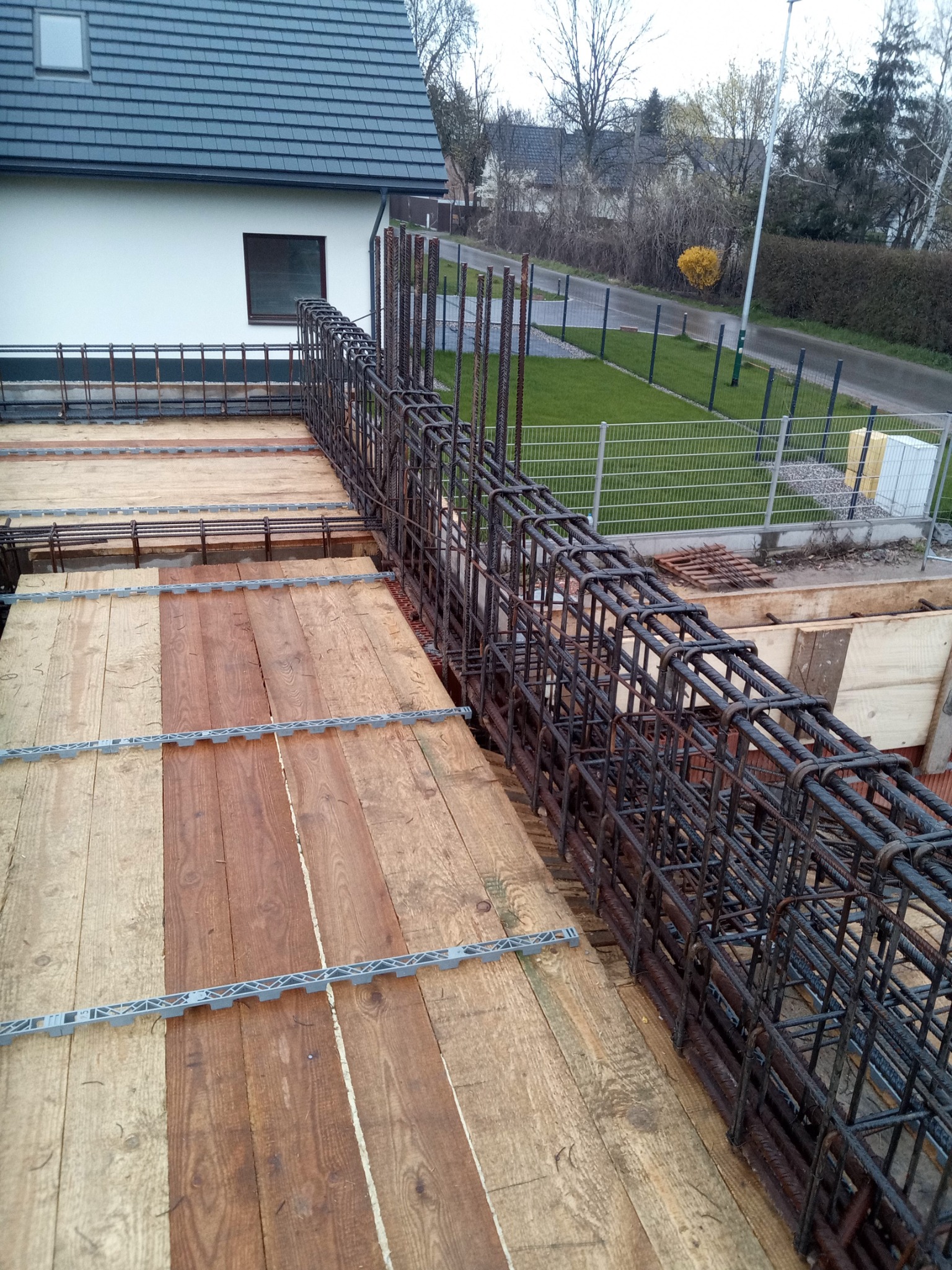 Zbrojenie pionowe z prętów stalowych w szalunku drewnianym na budowie domu, widok z góry na konstrukcję z elementami dystansowymi, w tle biały dom z szarą dachówką i zielony trawnik.