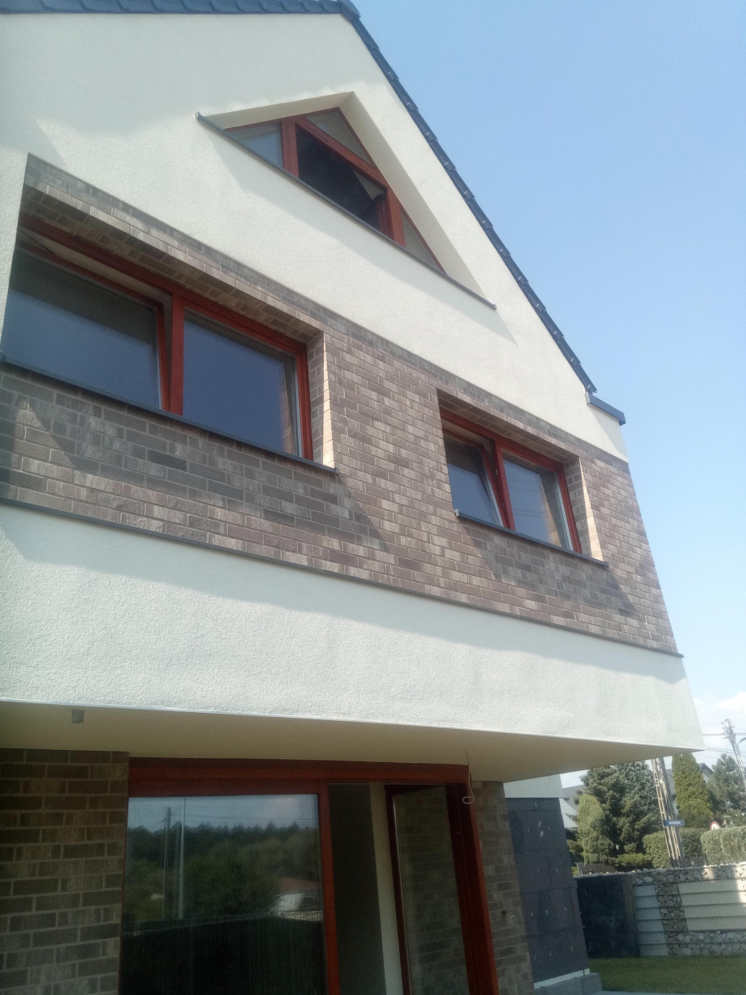 Nowoczesny dom z elewacją w odcieniach bieli i szarości, z fragmentem muru z cegły klinkierowej i oknami z ramami w kolorze mahoniu, widziany z dołu pod kątem.