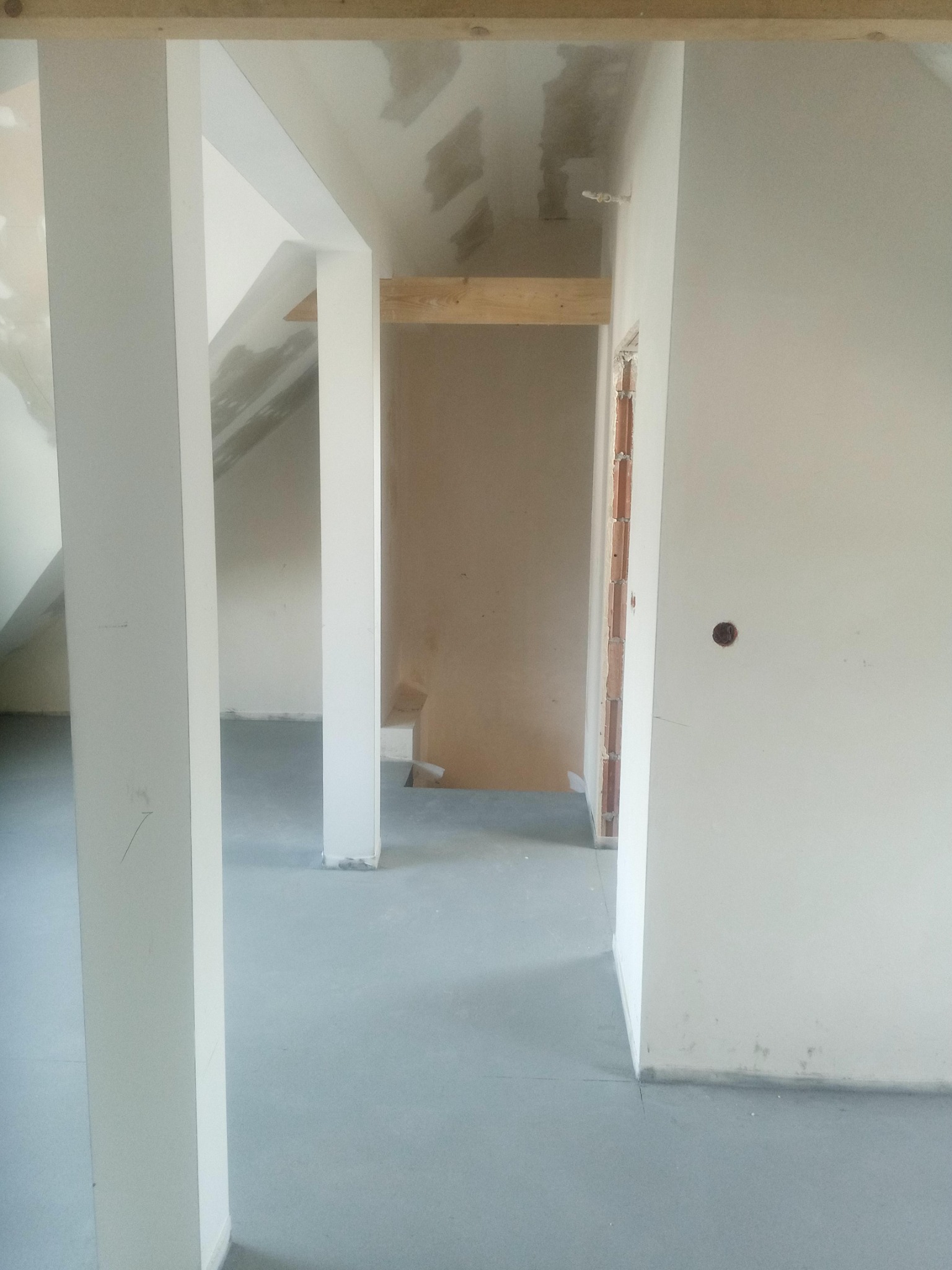 Wnętrze domu w trakcie budowy: surowe ściany, betonowa podłoga, drewniane elementy konstrukcyjne i widoczne cegły w otworze drzwiowym.