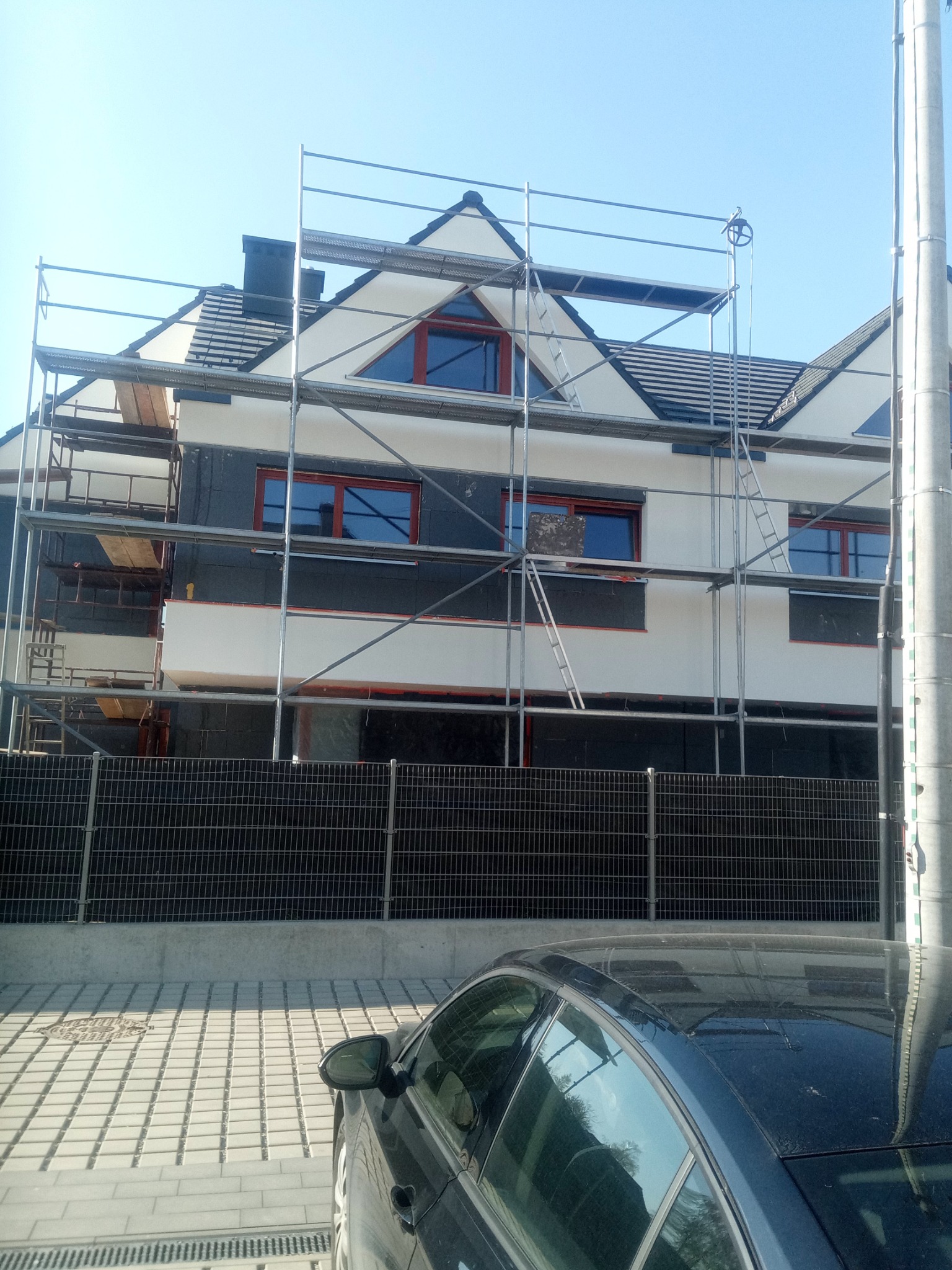 Dwupiętrowy dom w trakcie ocieplania, widoczne rusztowania, czarna izolacja termiczna na ścianach i częściowo nałożony biały tynk, brązowe ramy okienne, fragment zaparkowanego samochodu w dolnej...