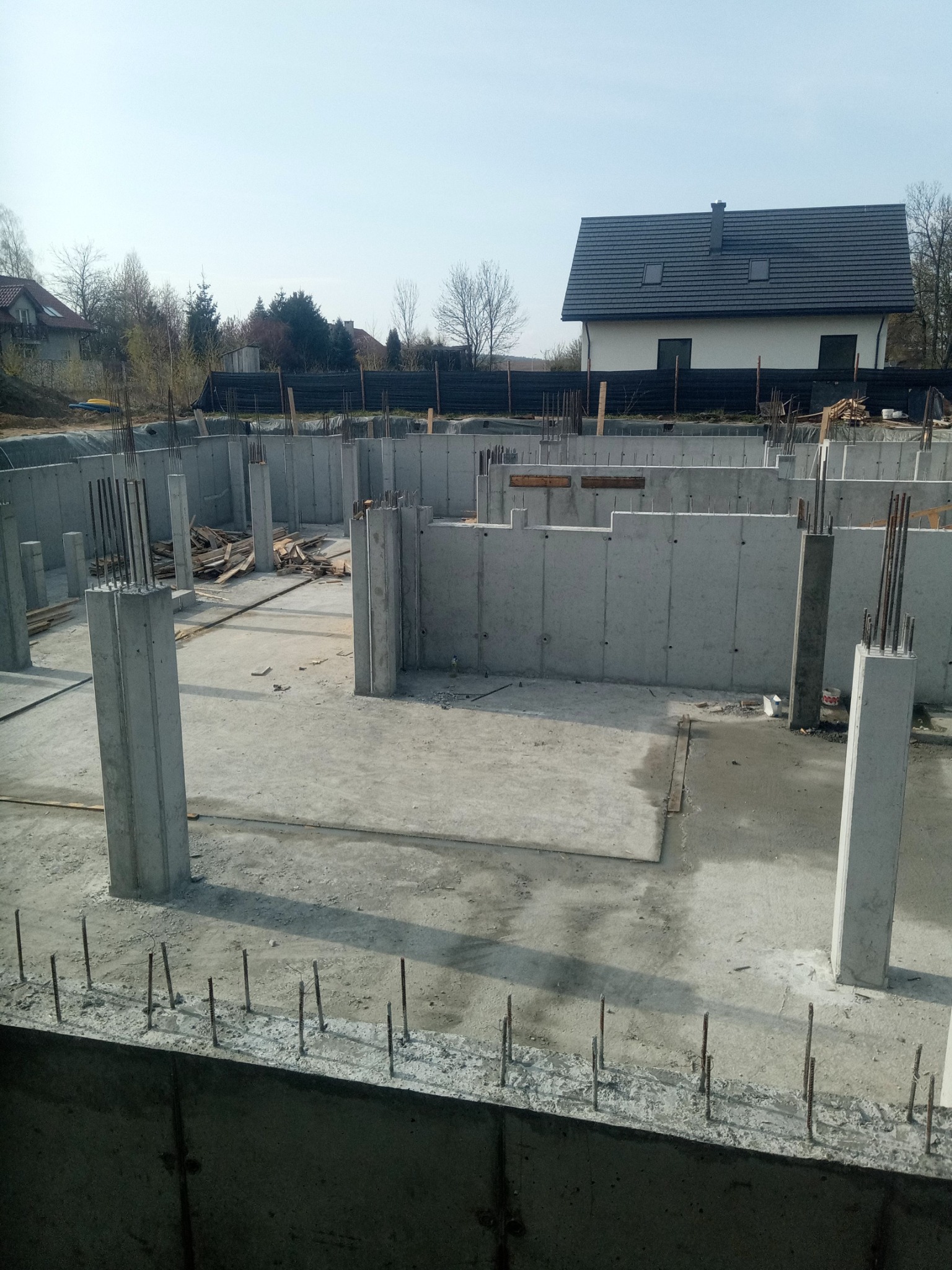 Surowe fundamenty domu z betonu, wystające pręty zbrojeniowe, widok na budynek mieszkalny w tle, wczesna faza budowy.