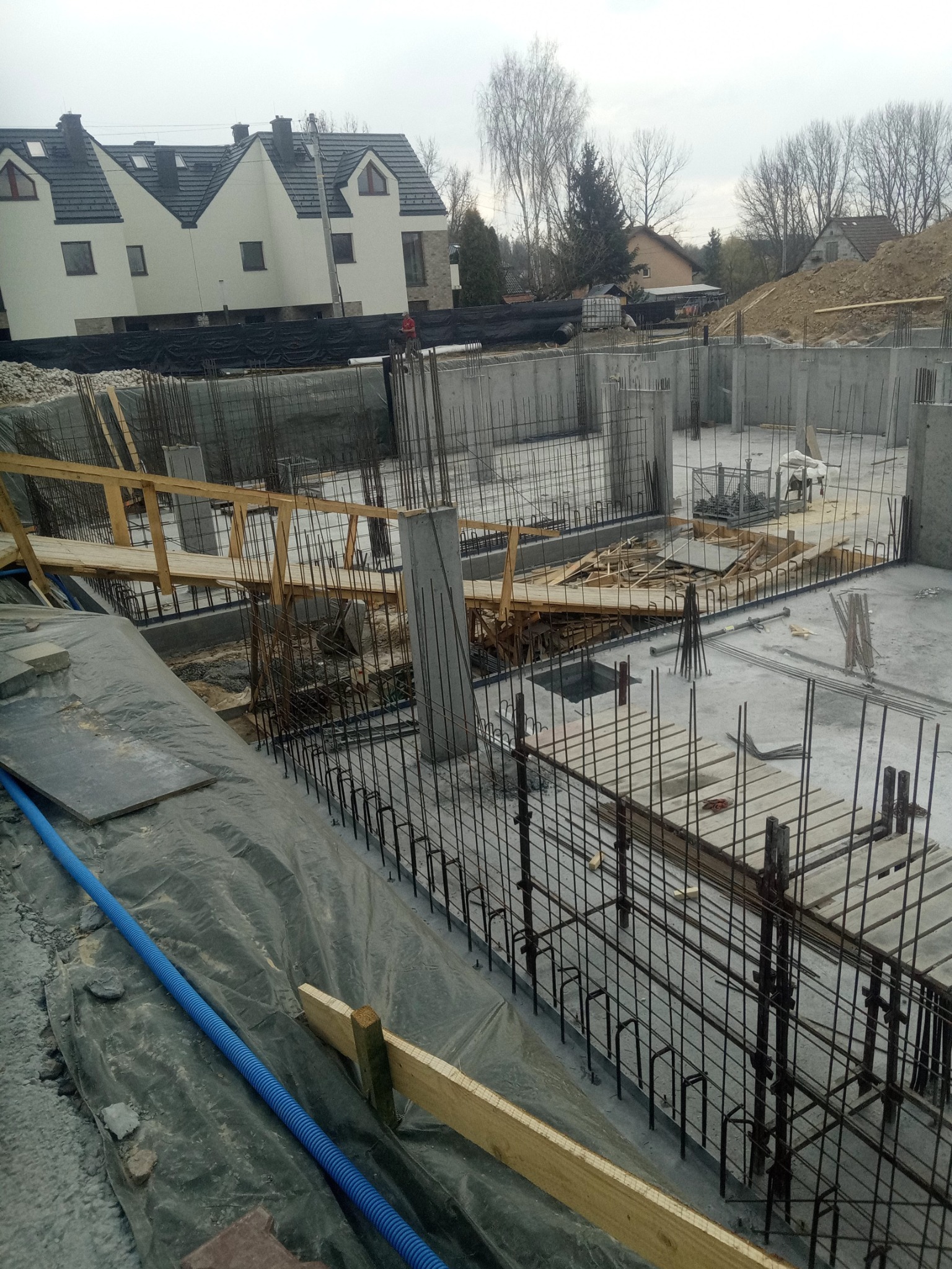Surowy stan fundamentów domu z widocznym zbrojeniem, szalunkami i elementami drewnianymi, na tle wzniesionego domu i pochmurnego nieba.