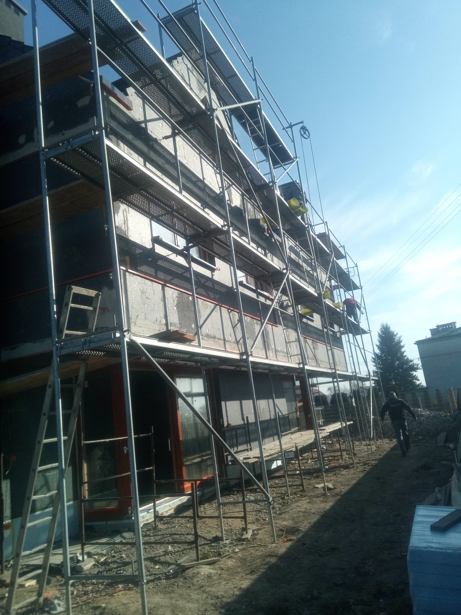 Budynek w trakcie ocieplania, widoczne rusztowania, częściowo ocieplona ściana, pracownicy na rusztowaniach, przygotowane materiały budowlane na ziemi.