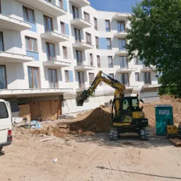 Żółta koparka Yanmar VIO80 przy wykopie pod fundamenty nowego apartamentowca z białymi balkonami, obok zaparkowany biały Volkswagen Transporter T4 i przenośna toaleta.