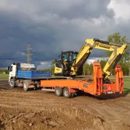 Żółta koparka Yanmar na pomarańczowej niskopodwoziowej przyczepie, transportowana przez ciężarówkę z niebieską skrzynią ładunkową, na tle pola i burzowego nieba.