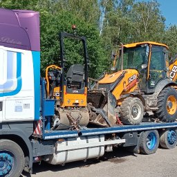 KRECIO - Ciężarówka DANN-TRANS LKW transportuje pomarańczową koparko-ładowarkę JCB i walec drogowego JCB na niskopodwoziowej platformie. W tle drzewa i budynek.