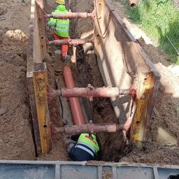 KRECIO - Wykop zabezpieczony szalunkiem stalowym. Wewnątrz pracownicy układają rury kanalizacyjne. Widoczna łyżka koparki i elementy infrastruktury.