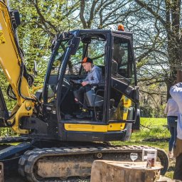 KRECIO - Żółto-czarna koparka Yanmar Vio80 z operatorem w kabinie, na tle zieleni w Kościerzynie. Widoczny fragment naczepy i osoby obserwujące pracę.
