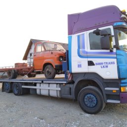 KRECIO - Laweta z pomarańczowym, zabytkowym samochodem na platformie, zaparkowana na żwirowym podłożu. Widoczny fragment budynku w tle i osoba na rowerze.