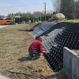 KRECIO - Pracownik w czerwonej koszuli wypełnia geokratę kamieniami na zboczu. W tle widoczny sprzęt budowlany i domy. Słoneczny dzień, budowa w toku.