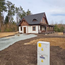 KRECIO - Ukończony dom z podjazdem z kamienia, wyrównany teren wokół. Widoczna skrzynka elektryczna i osoba w oddali. Prace ziemne w toku, teren przygotowany pod trawnik.