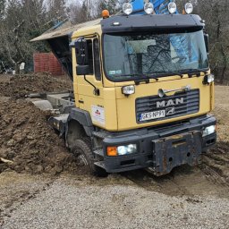 KRECIO - Żółta ciężarówka MAN w trakcie prac ziemnych na działce. Widoczna sterta ziemi i pracownik z łopatą. Działka z drzewami w tle. Tablica rejestracyjna GKS NP91.