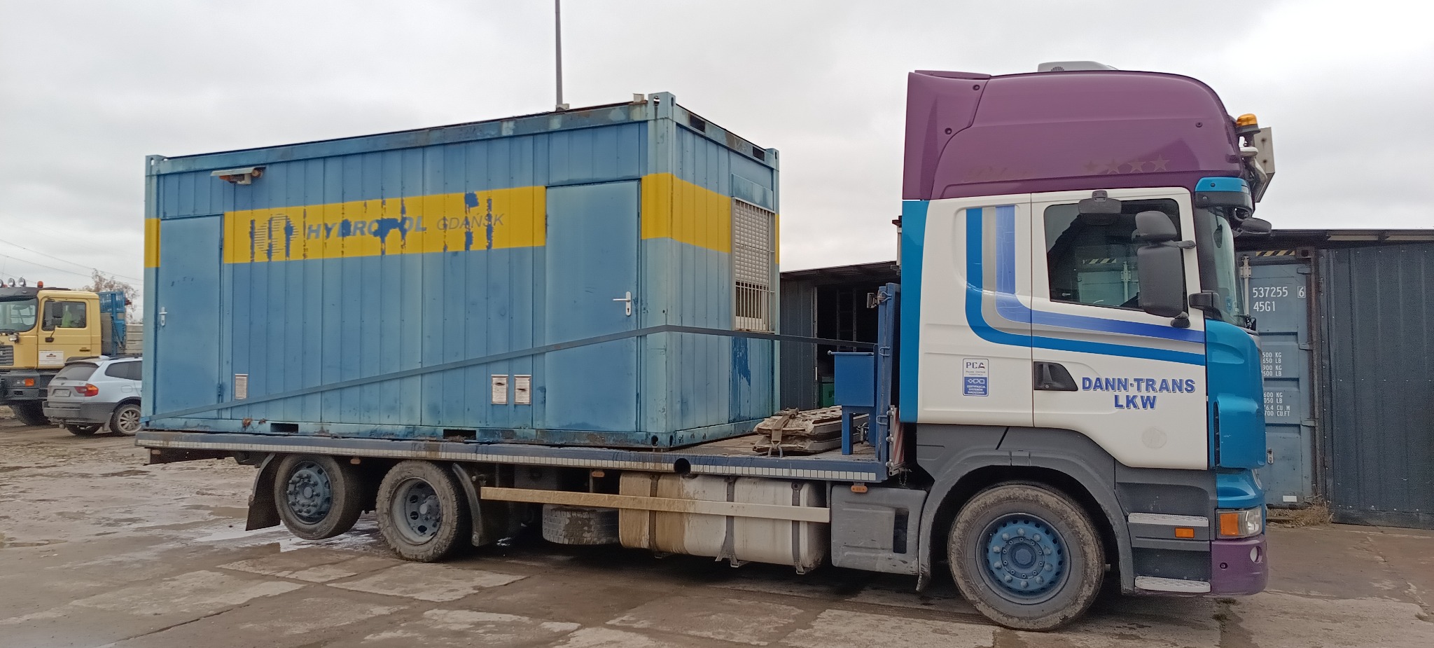 Ciężarówka z niebieskim kontenerem z żółtym pasem, na którym widnieje napis 'HYDROL GDAŃSK', zaparkowana na betonowym placu. Widoczny napis 'DANN-TRANS LKW' na kabinie.