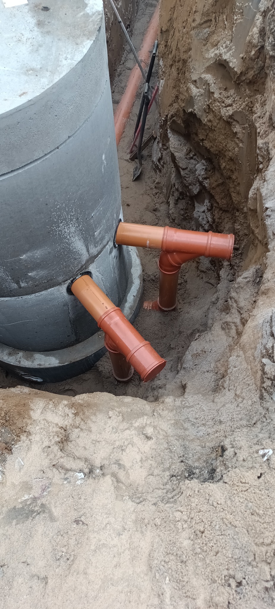 Betonowy zbiornik z podłączonymi rurami kanalizacyjnymi w wykopie. Widoczne narzędzia i piasek. Instalacja szamba betonowego w trakcie montażu.