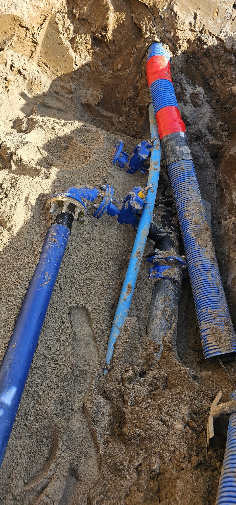 Niebieskie rury z zaworami w piasku wykopu, jedna z rur owinięta czerwoną taśmą. Widok z góry na instalację wodno-kanalizacyjną w trakcie prac ziemnych.