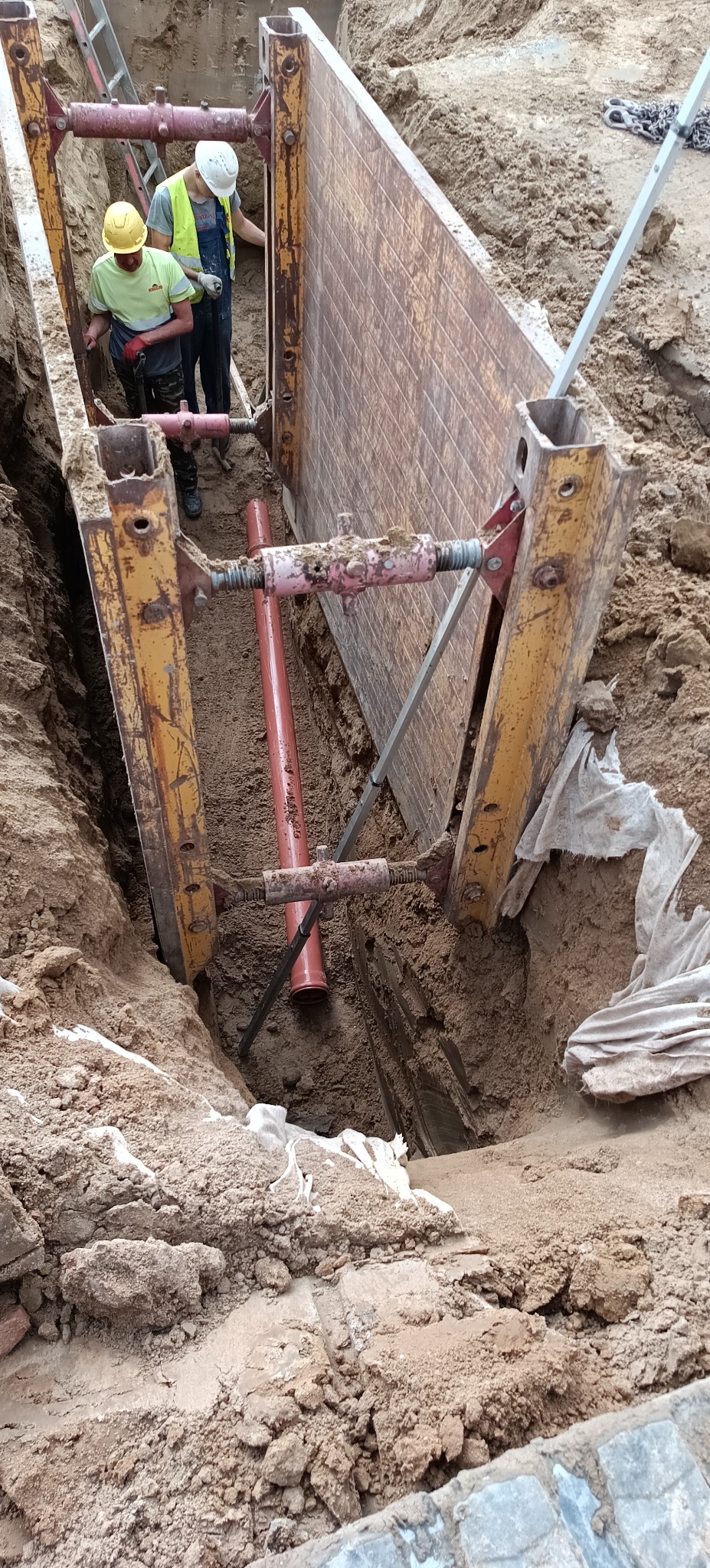 Głęboki wykop zabezpieczony szalunkiem stalowym, wewnątrz rura kanalizacyjna. Dwóch pracowników w kaskach nadzoruje prace. Widoczne ślady ziemi na elementach konstrukcji.