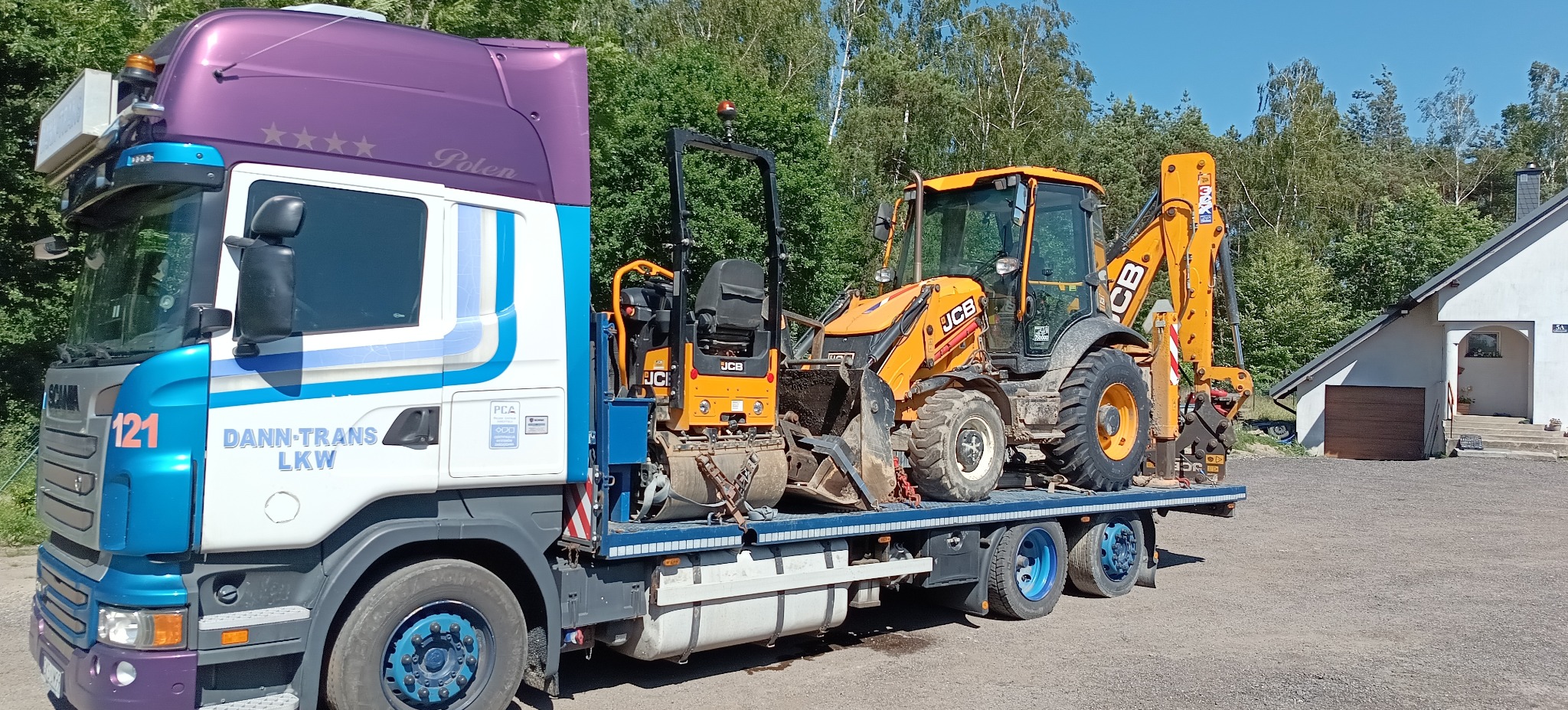Ciężarówka DANN-TRANS LKW transportuje pomarańczową koparko-ładowarkę JCB i walec drogowego JCB na niskopodwoziowej platformie. W tle drzewa i budynek.
