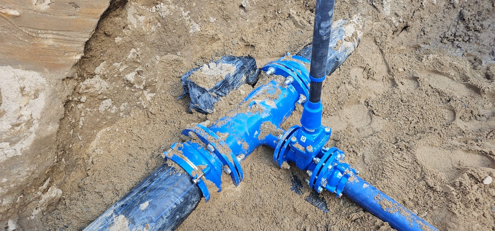 Niebieskie rury złączone w ziemi, widoczne ślady piasku. Instalacja wodno-kanalizacyjna w trakcie montażu, złącza skręcone śrubami. Widok z góry.
