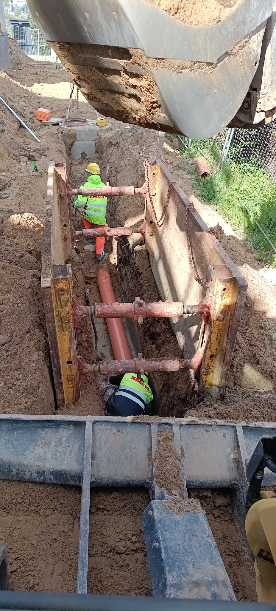 Wykop zabezpieczony szalunkiem stalowym. Wewnątrz pracownicy układają rury kanalizacyjne. Widoczna łyżka koparki i elementy infrastruktury.