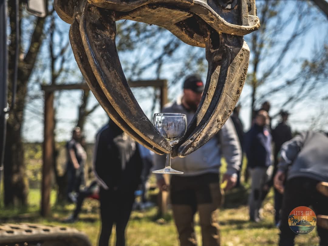 Kieliszek wina trzymany przez ramię koparki na tle grupy osób i zieleni. Nietypowe ujęcie z perspektywy maszyny budowlanej, podkreślające kontrast i zabawę.