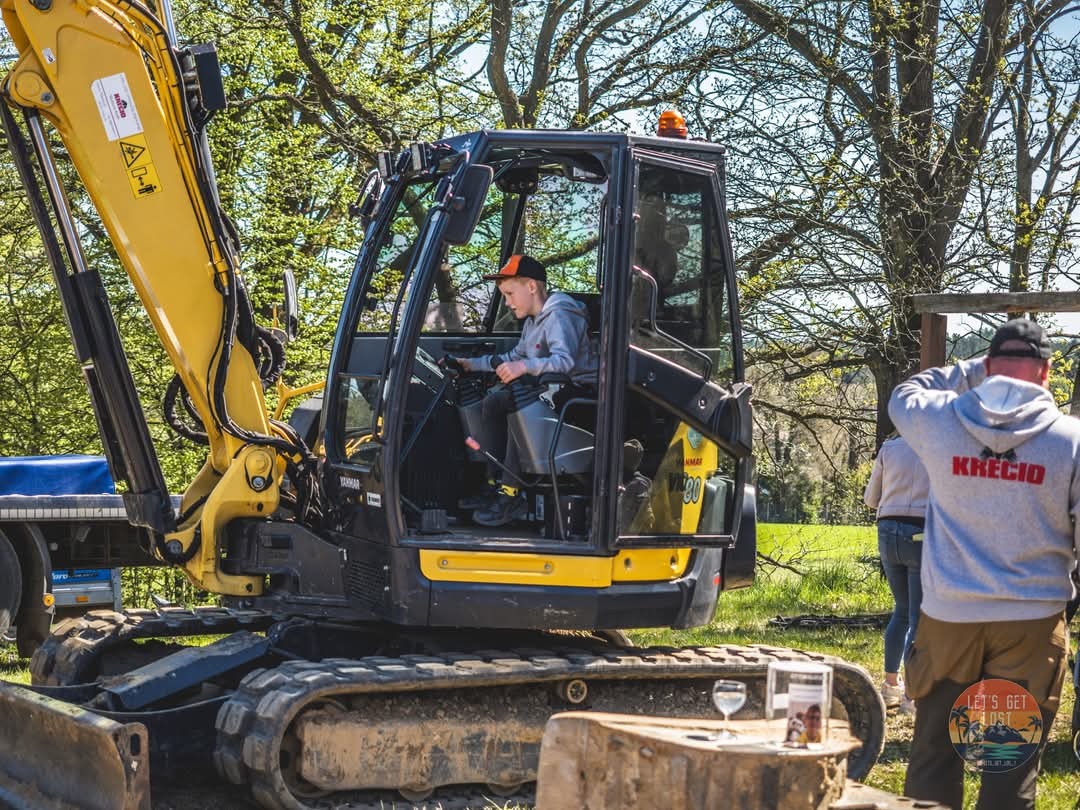 Żółto-czarna koparka Yanmar Vio80 z operatorem w kabinie, na tle zieleni w Kościerzynie. Widoczny fragment naczepy i osoby obserwujące pracę.