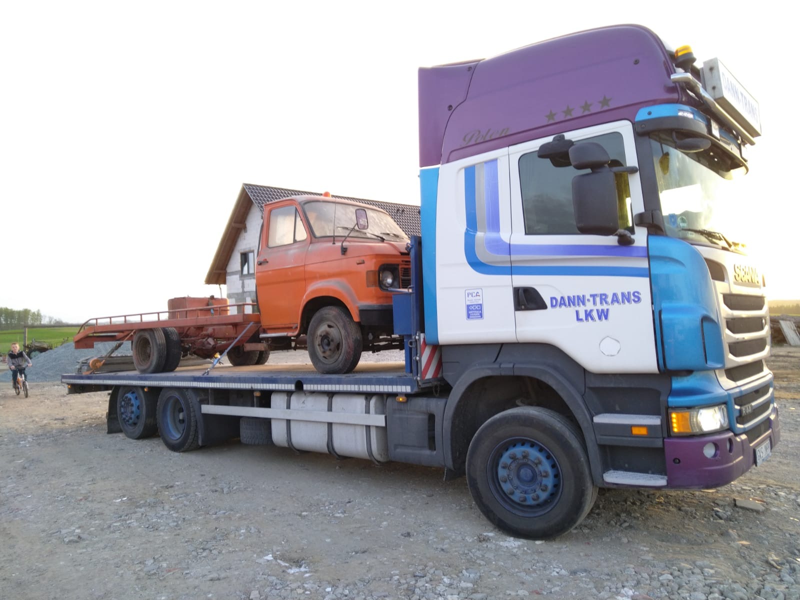 Laweta z pomarańczowym, zabytkowym samochodem na platformie, zaparkowana na żwirowym podłożu. Widoczny fragment budynku w tle i osoba na rowerze.