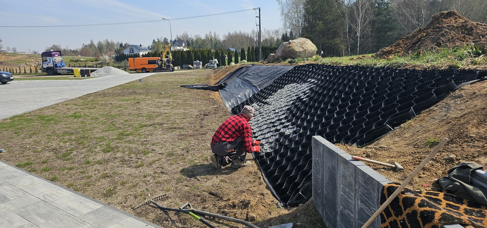 Pracownik w czerwonej koszuli wypełnia geokratę kamieniami na zboczu. W tle widoczny sprzęt budowlany i domy. Słoneczny dzień, budowa w toku.