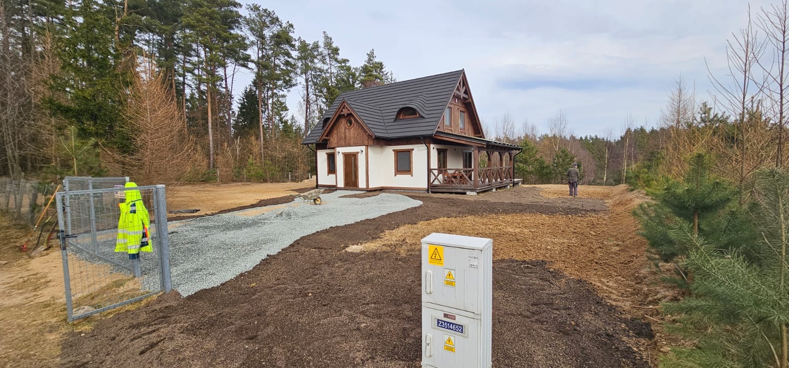Ukończony dom z podjazdem z kamienia, wyrównany teren wokół. Widoczna skrzynka elektryczna i osoba w oddali. Prace ziemne w toku, teren przygotowany pod trawnik.