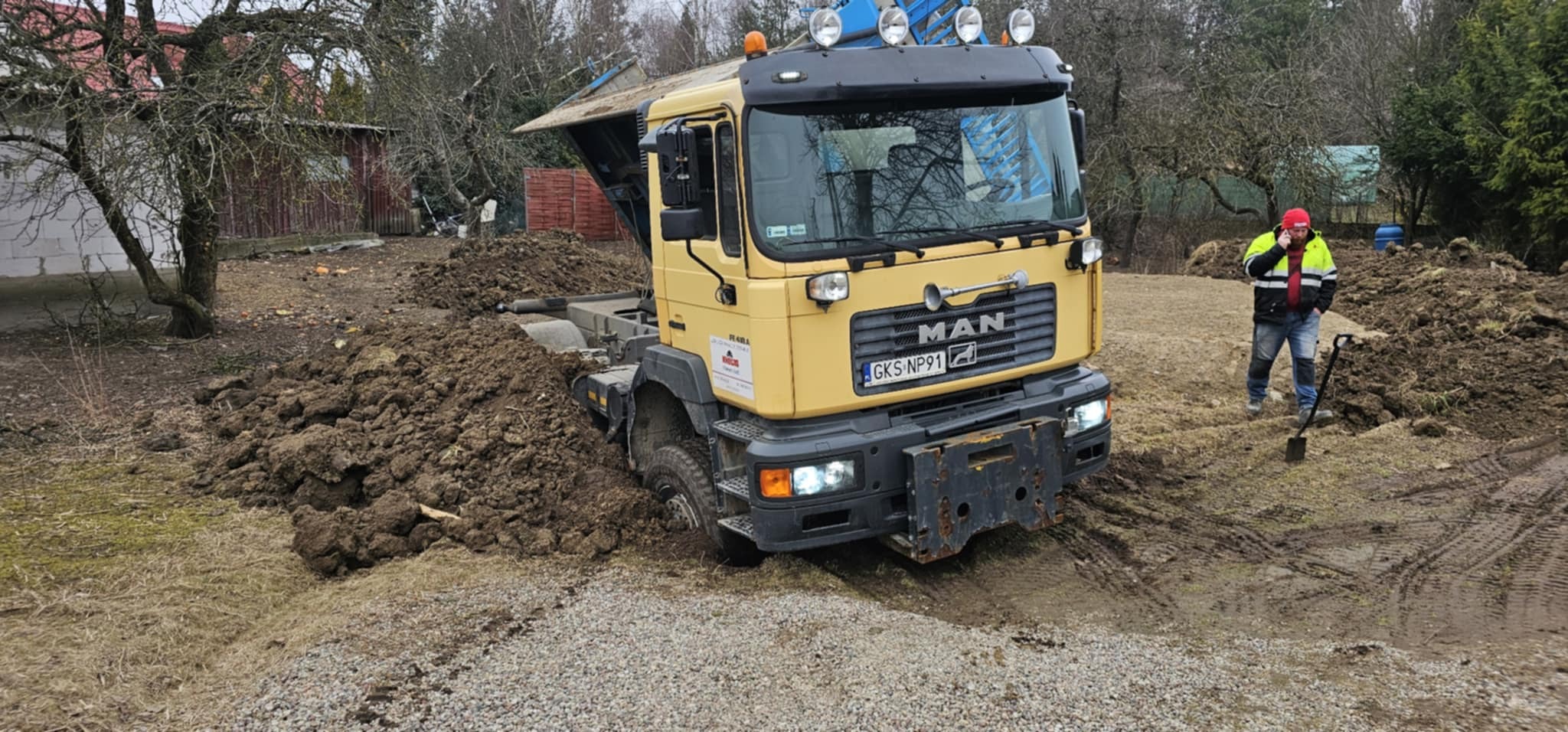 Żółta ciężarówka MAN w trakcie prac ziemnych na działce. Widoczna sterta ziemi i pracownik z łopatą. Działka z drzewami w tle. Tablica rejestracyjna GKS NP91.