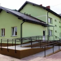 Jasnozielony budynek z ciemnobrązowym dachem, rynnami i podmurówką, z rampą dla wózków inwalidzkich prowadzącą do wejścia. Widoczne okna z białymi ramami.