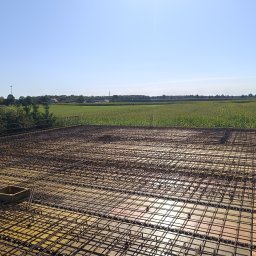 ZAKŁAD REMONTOWO-BUDOWLANY ANTONI SZNEL - Zbrojenie stropu domu jednorodzinnego, widok z góry na siatkę z prętów stalowych ułożoną na żółtych deskach szalunkowych. W tle zielone pole i błękitne niebo.