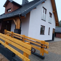 ZAKŁAD REMONTOWO-BUDOWLANY ANTONI SZNEL - Nowoczesny dom jednorodzinny z podjazdem dla osób z niepełnosprawnościami. Biała elewacja, drewniane elementy wykończeniowe, czarny dach. Widok z zewnątrz.