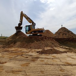 ZAKŁAD REMONTOWO-BUDOWLANY ANTONI SZNEL - Żółta koparka CAT M315D nabiera piasek na tle błękitnego nieba z chmurami. Widoczne ślady opon na piasku i wieża telekomunikacyjna na szczycie piaskowej góry.