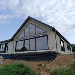 ZAKŁAD REMONTOWO-BUDOWLANY ANTONI SZNEL - Niedokończony dom z białych bloczków, z dużymi oknami i czarną stolarką, na tle pochmurnego nieba. Widoczne prace ziemne wokół budynku.