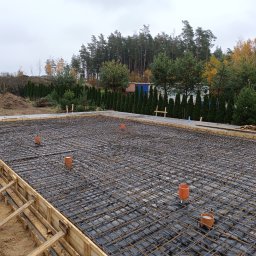 ZAKŁAD REMONTOWO-BUDOWLANY ANTONI SZNEL - Zbrojenie fundamentów pod budowę domu jednorodzinnego. Widoczne stalowe pręty tworzące siatkę, szalunek z desek i pionowe rury kanalizacyjne. W tle drzewa i pochmurne niebo.