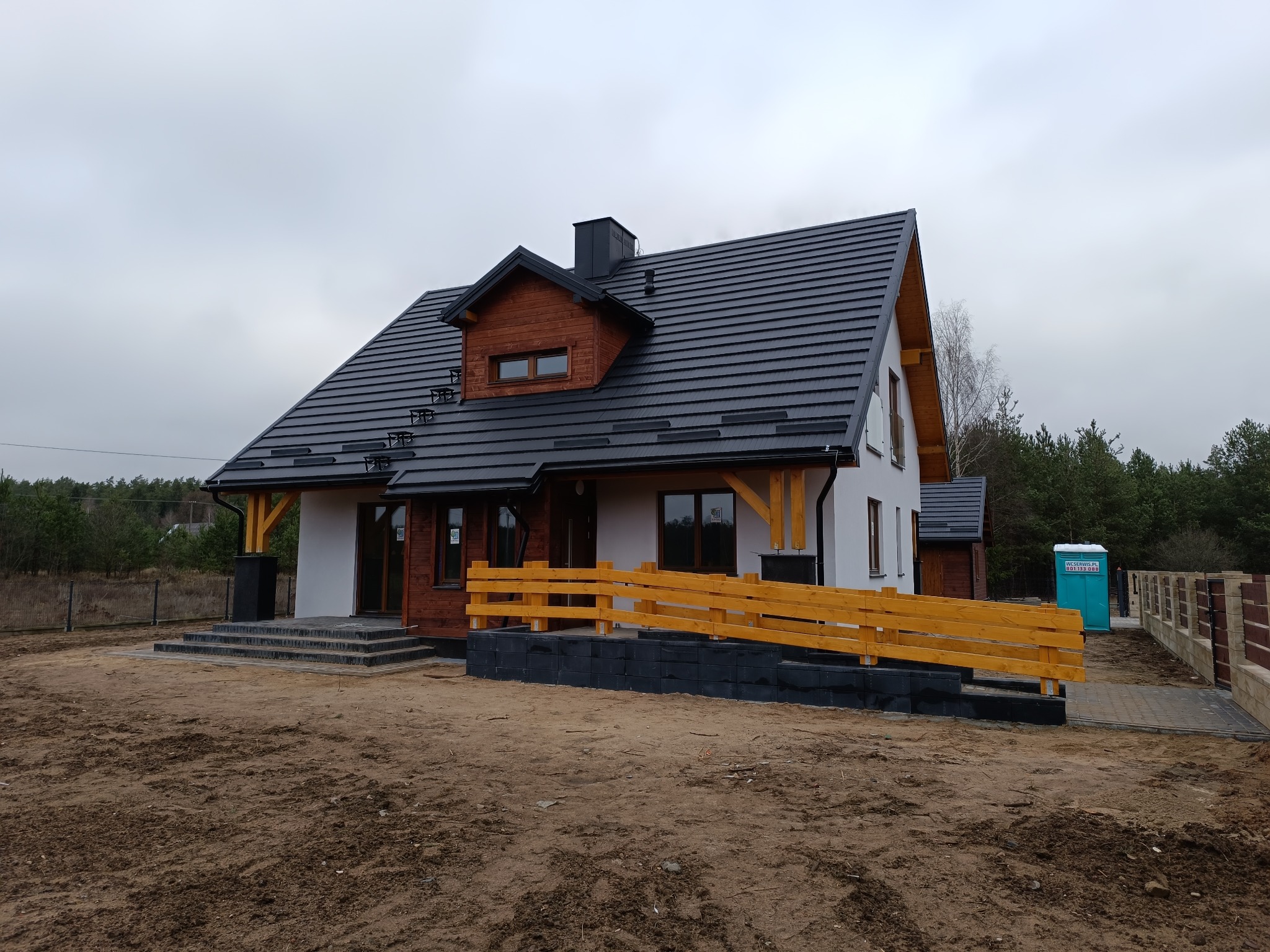 Niedawno wybudowany dom jednorodzinny z czarną dachówką, drewnianymi elementami konstrukcyjnymi oraz żółtą barierką, na działce z widoczną ziemią i przenośną toaletą w tle.
