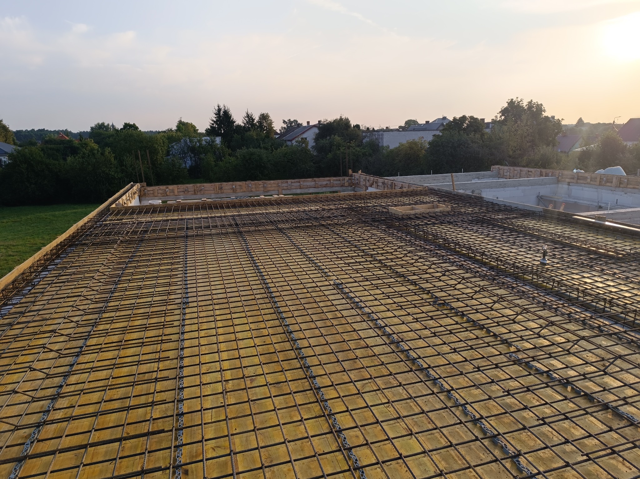 Zbrojenie płyty fundamentowej na deskowaniu z widocznymi elementami dystansowymi, w tle zieleń i zabudowa jednorodzinna oświetlona zachodzącym słońcem.