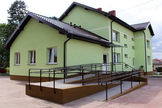 Jasnozielony budynek z ciemnobrązowym dachem, rynnami i podmurówką, z rampą dla wózków inwalidzkich prowadzącą do wejścia. Widoczne okna z białymi ramami.