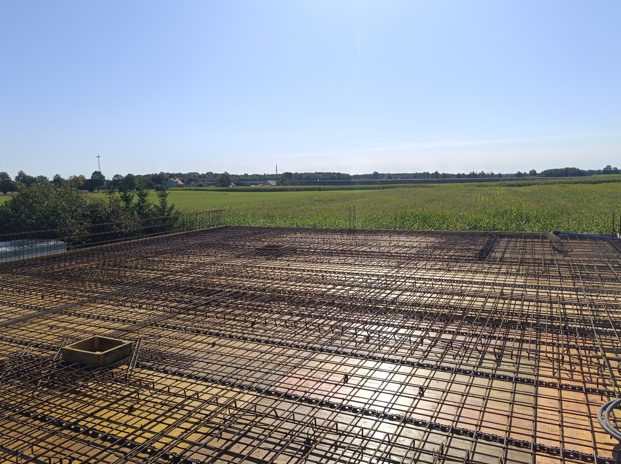 Zbrojenie stropu domu jednorodzinnego, widok z góry na siatkę z prętów stalowych ułożoną na żółtych deskach szalunkowych. W tle zielone pole i błękitne niebo.