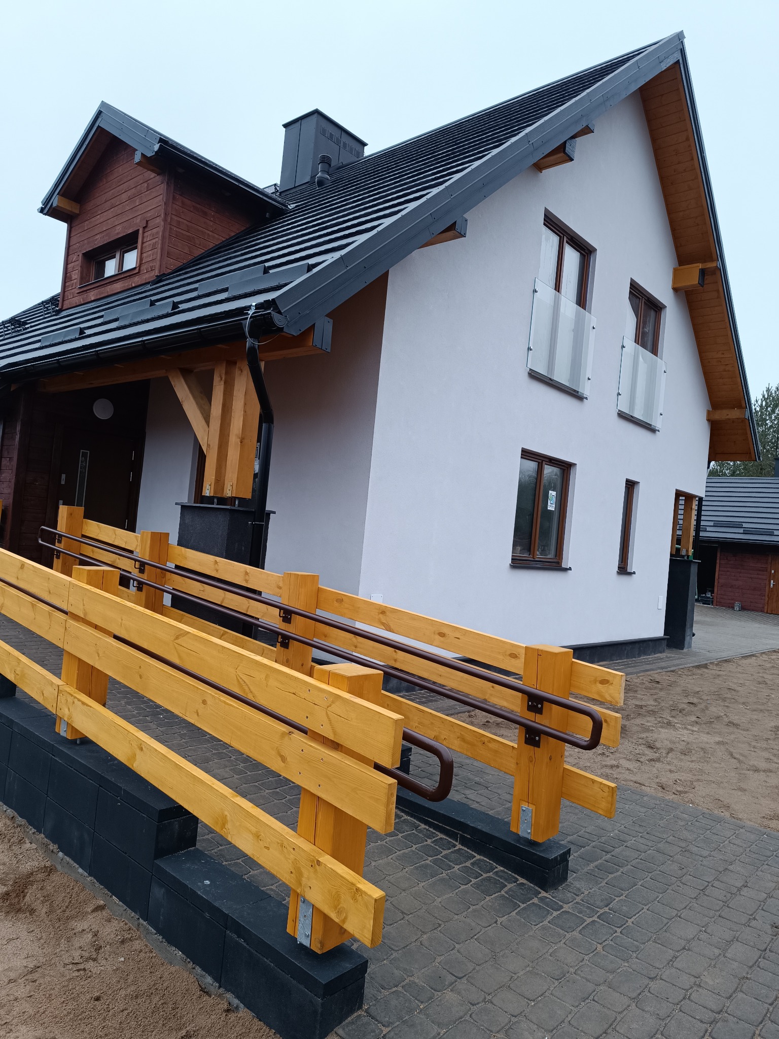 Nowoczesny dom jednorodzinny z podjazdem dla osób z niepełnosprawnościami. Biała elewacja, drewniane elementy wykończeniowe, czarny dach. Widok z zewnątrz.