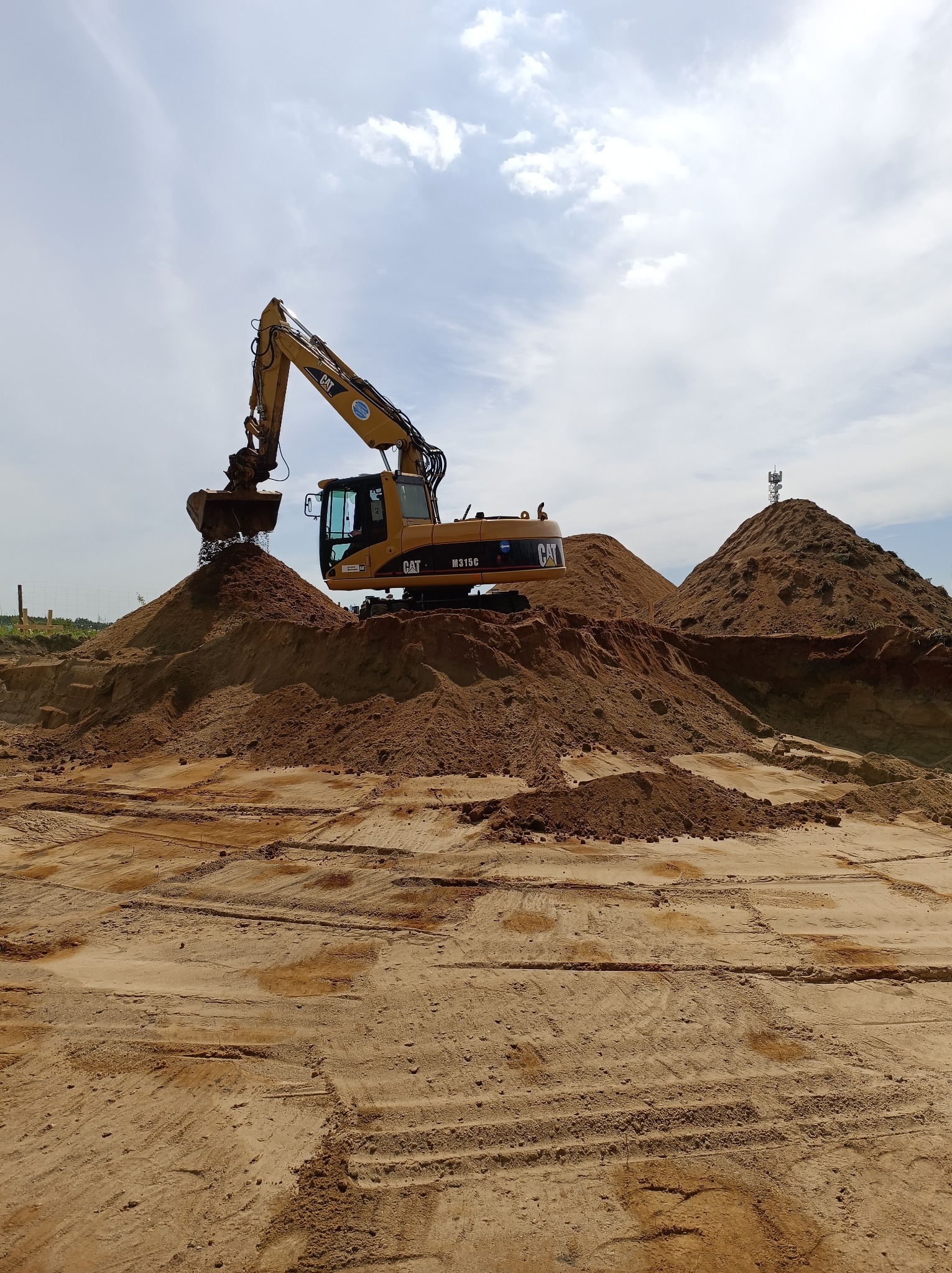 Żółta koparka CAT M315D nabiera piasek na tle błękitnego nieba z chmurami. Widoczne ślady opon na piasku i wieża telekomunikacyjna na szczycie piaskowej góry.