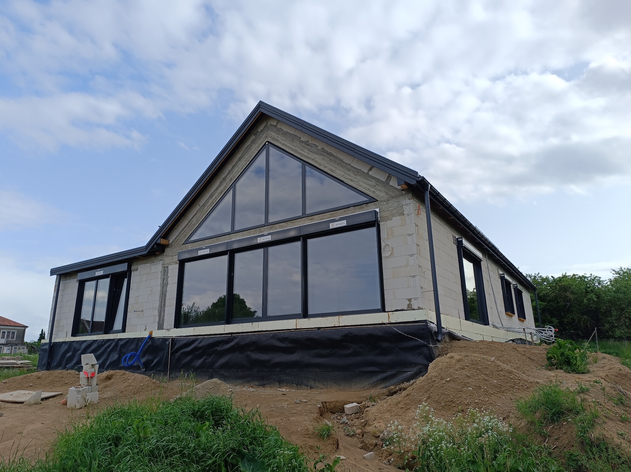 Niedokończony dom z białych bloczków, z dużymi oknami i czarną stolarką, na tle pochmurnego nieba. Widoczne prace ziemne wokół budynku.