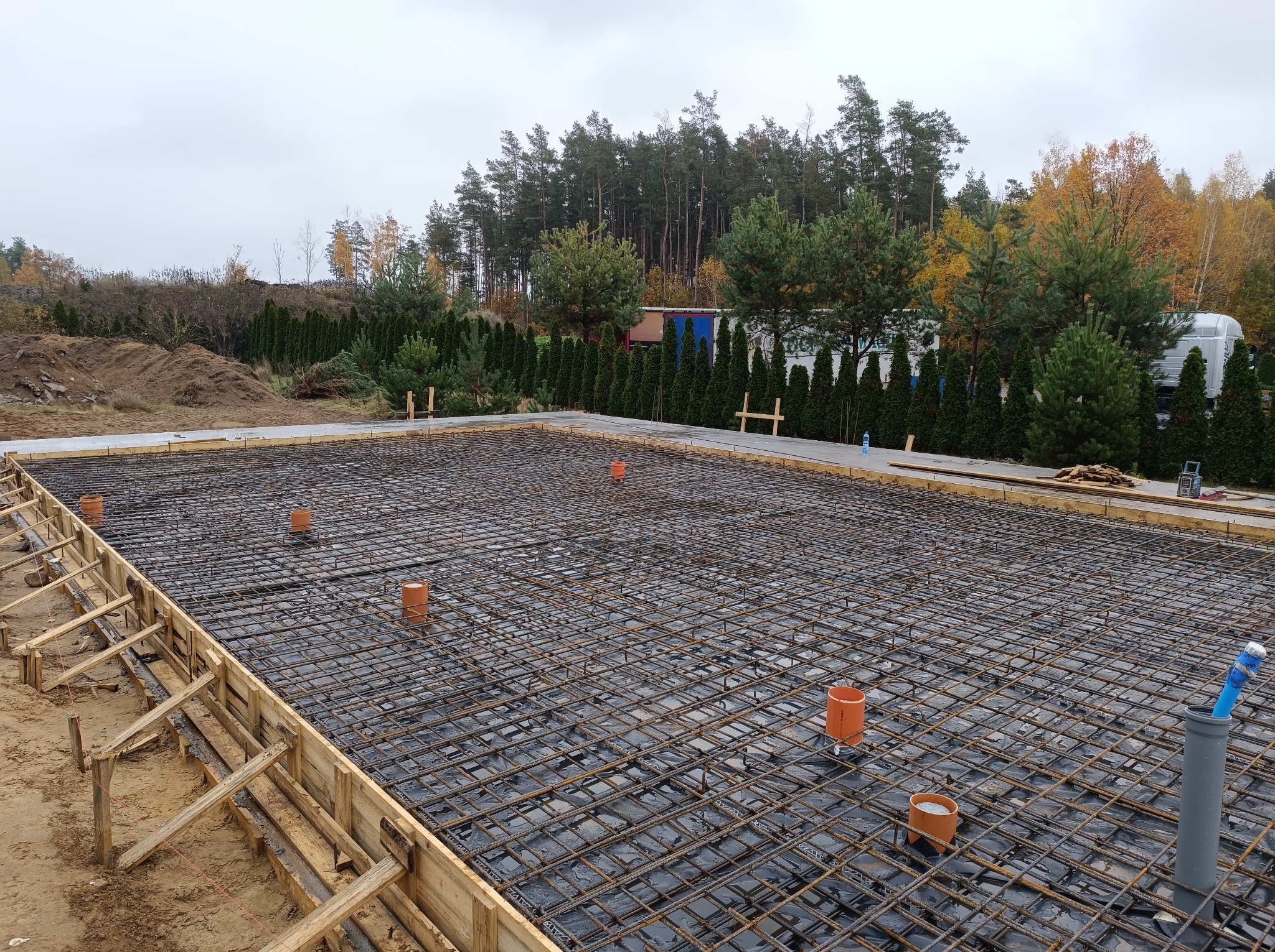 Zbrojenie fundamentów pod budowę domu jednorodzinnego. Widoczne stalowe pręty tworzące siatkę, szalunek z desek i pionowe rury kanalizacyjne. W tle drzewa i pochmurne niebo.