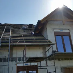 Budowa dachu z częściowo ułożoną dachówką, widoczna drabina i rusztowanie przy budynku z pustaków. Słońce w kadrze.