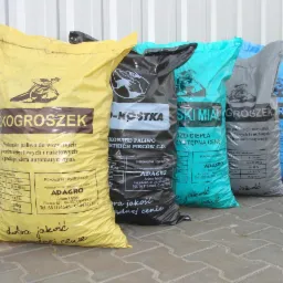 Rząd worków z ekogroszkiem i innymi rodzajami węgla, różnych kolorów, z widocznymi etykietami i logotypem firmy Adagro, ustawionych na zewnątrz budynku.