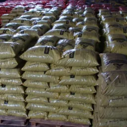 Magazyn ekogroszku: rzędy worków ułożonych na paletach, z widocznymi etykietami 'EKOGROSZEK' i oznaczeniami producenta. Czerwone worki w tle.