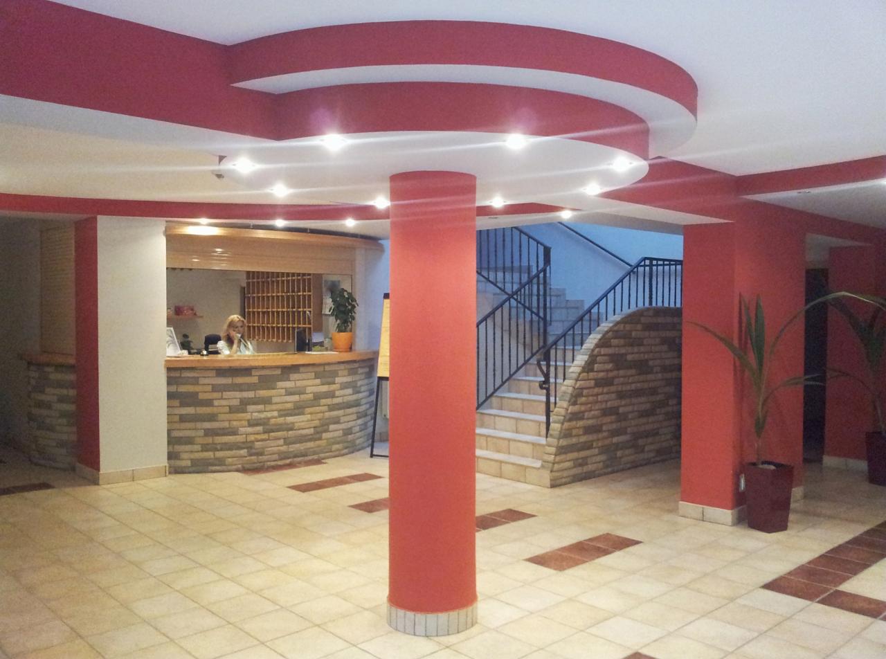 Recepcja w hotelu z widoczną pracownicą za kamienną ladą, kolumną w kolorze czerwonym i schodami prowadzącymi na wyższe piętro.
