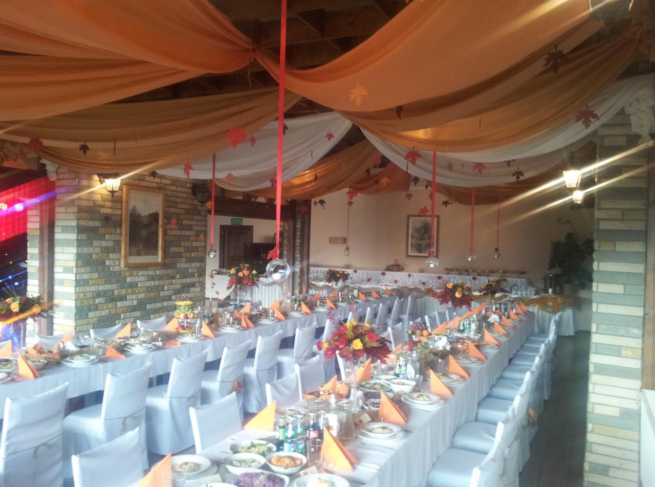 Sala bankietowa udekorowana na jesienną uroczystość: białe obrusy, pomarańczowe akcenty, dekoracje z liści i zwisające wstążki.