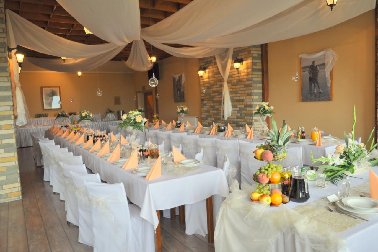 Sala bankietowa z długimi stołami, nakryta na przyjęcie weselne. Białe obrusy, pomarańczowe serwetki, owoce i kwiaty. Dekoracja sufitu z białych tkanin.