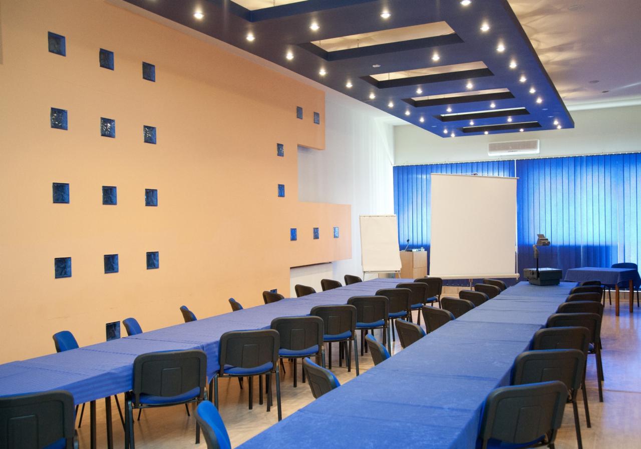 Sala konferencyjna z długimi stołami przykrytymi niebieskimi obrusami, krzesłami, ekranem projekcyjnym i projektorem. Dekoracyjna ściana z niebieskimi elementami.